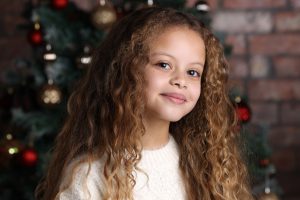 Christmas Photoshoots. Image of a young girl in a white jumper with a Christmas tree behind her